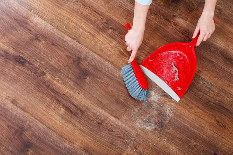 A closeup photo of a woman holding a dustpan and brush, sweeping dust and dirt off a wooden floor.
