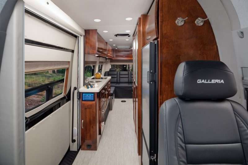 Interior of a Coachmen Galleria camper van for sale, shown from the driver's seat looking back. There are beautiful hardwood cabinets in a cherry finish lining the cab of the coach. Two coat hooks are located behind the black leather captain's chair. A galley kitchen is located on the passenger side of the RV. At the very back of the coach, a black leather sofa is visible.