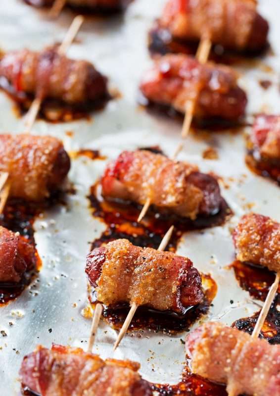 Little Smokie sausages wrapped in bacon and a brown sugar glaze are arranged on a foil-lined pan. The bacon is held in place with toothpicks.