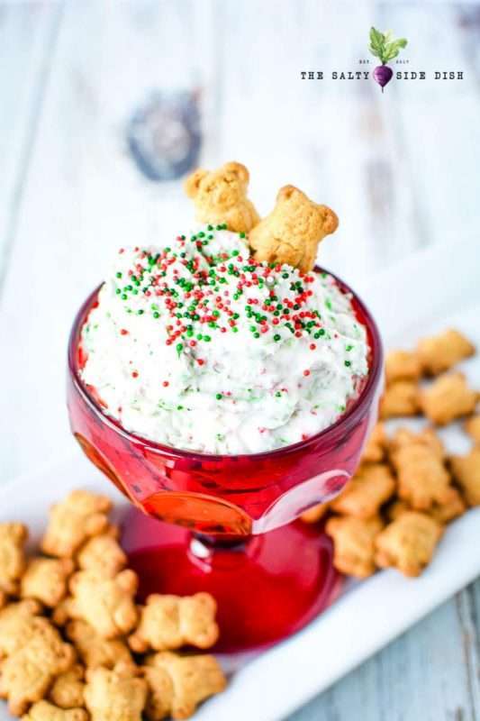 A red glass with a large opening is filled with fluffy white Christmas Cookie Dip. This easy holiday dip recipe is served with teddy graham crackers and red and green sprinkles. 