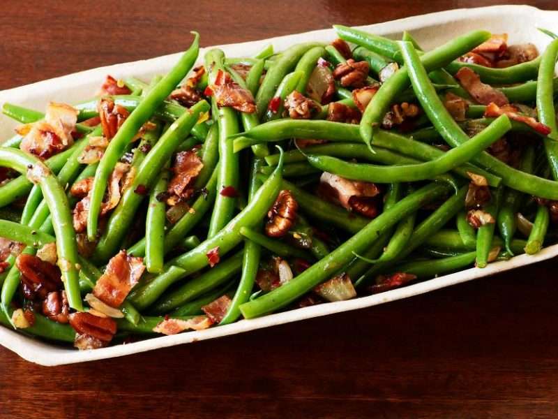 A white serving platter is filled with cooked green beans and bacon. Chopped pecans are added to top off this easy holiday side dish.