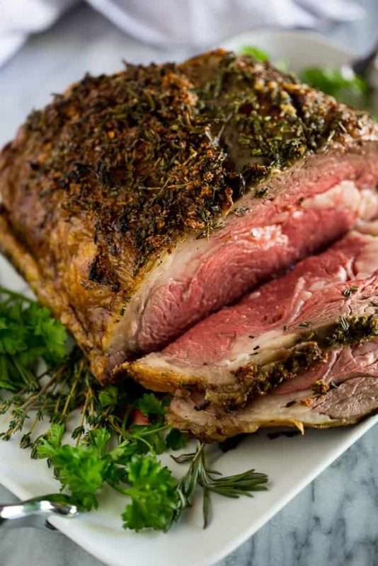 A prime rib with a crispy brown, herb crust sits on a white plate on a marble counter top.