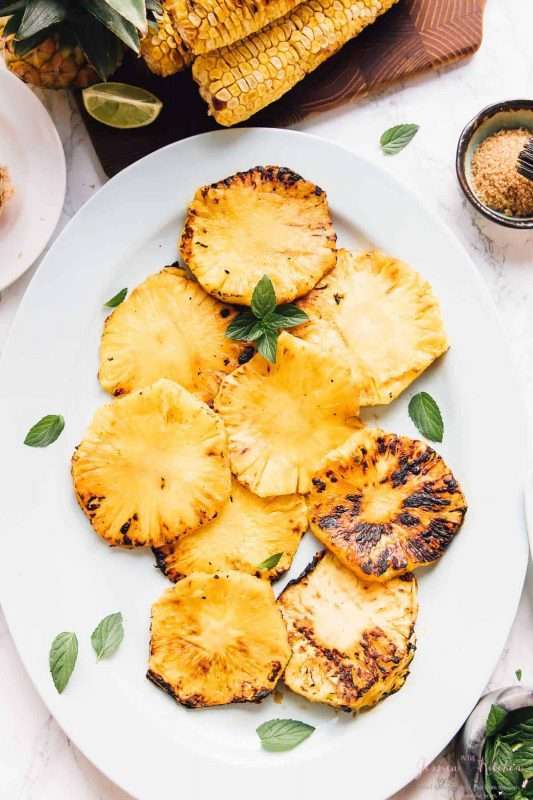 Thick slices of grilled pineapple are arranged on a white serving plate. The edges of the pineapple have caramelized to a deep brown-black color.  