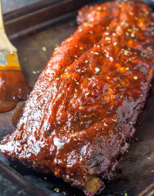 A rack of glazed ribs sits on a sheet pan. A basting brush with barbecue sauce is visible in the corner of the photo. These ribs were made in an oven for an easy holiday main course recipe.