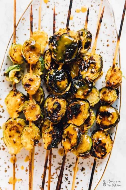 Several charred skewers of grilled Brussels sprouts are arranged on a long serving plate speckled with color.