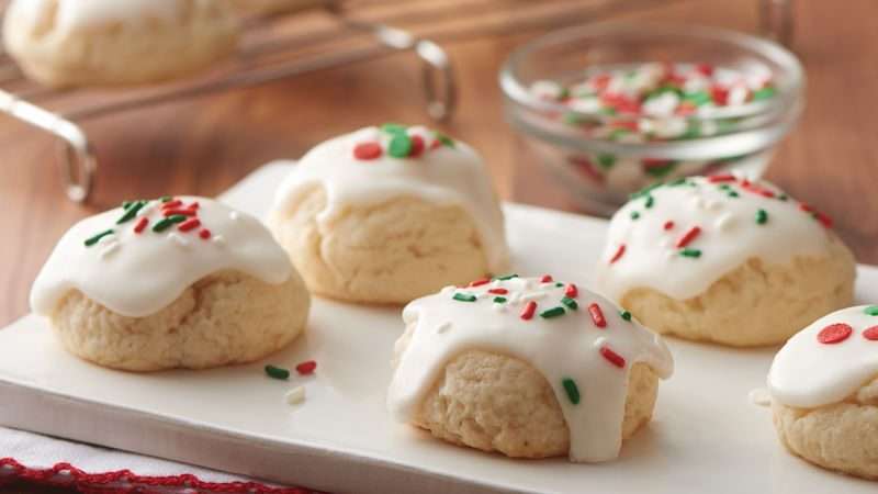 Round sugar cookies topped with white icing and festive red, green, and white sprinkles are arranged on a white slab. In the background, a cookie cools on a rack and a small bowl of extra sprinkles sits next to it.