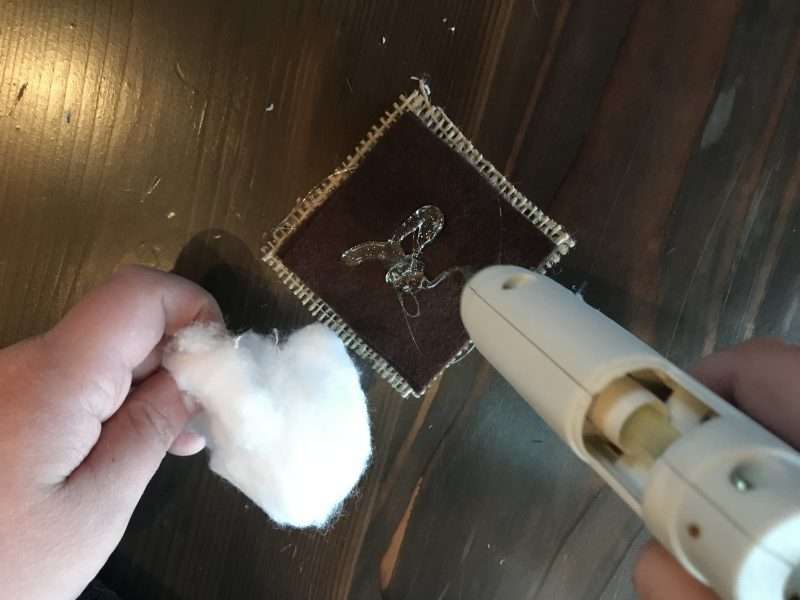 A dab of hot glue is added to the center of a square of brown felt sitting on top of a piece of burlap. The person assembling the S'mores ornament is holding a fluffed cotton ball in their left hand and a glue gun in their right hand.