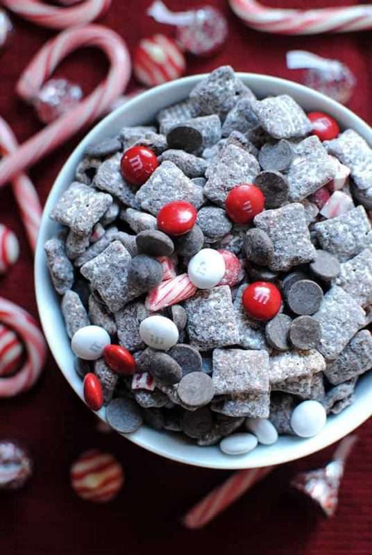 A white bowl is filled with Peppermint Bark Puppy Chow. Candy canes, peppermint striped Hershey's kisses, and other red and white candies surround the bowl in the background. 