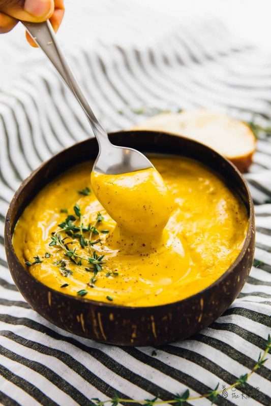 A spoon dives into a bowl of Roasted Sweet Potato Soup served with a slice of thick white bread. The bowl sits on a black and white striped tablecloth. This easy holiday recipe uses seasonal vegetables for a flavorful soup. 