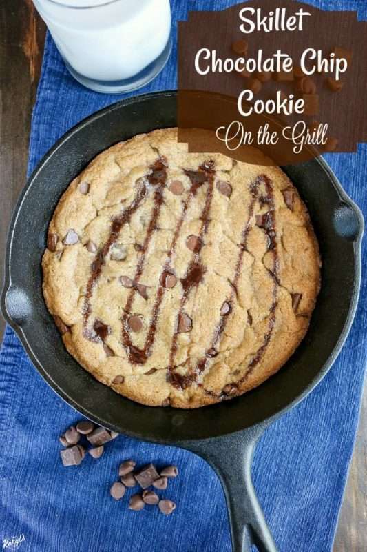 A large cast iron skillet is filled with a chocolate chip cookie. The cookie has browned edges and is topped with a swirl of chocolate sauce. The background is a wooden table with denim fabric and chocolate chips and chocolate chunks nearby.