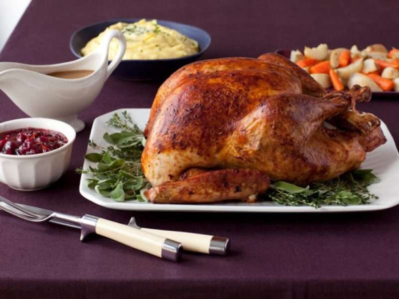 A browned thanksgiving turkey is plated on a maroon tablecloth to illustrate the results of this simple holiday recipe. To the left of the turkey is a small bowl of cranberry jelly and a gravy boat filled with brown gravy. In the background are a bowl of creamy mashed potatoes and a bowl of baby carrots and sliced potatoes.