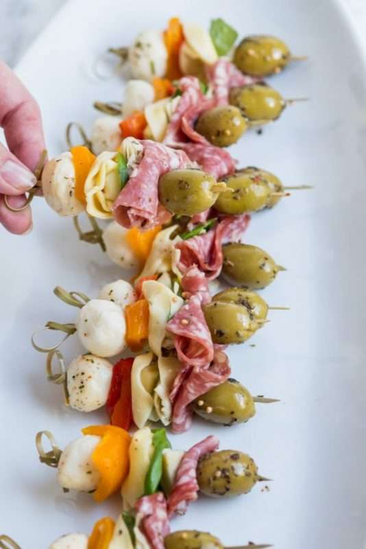 A hand holds a miniature skewer loaded with various goodies over a plateful of the prepared skewers. They include mozzarella balls, sliced sweet peppers, tortellini pasta, folded salami slices, and green olives. 