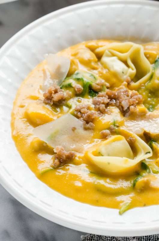 A white bowl is filled with Butternut Squash, Sausage, and Tortelloni Soup. The soup is a bright yellow-orange color and topped with pale Parmesan cheese, brown sausage crumbles, and a hint of green garnish.