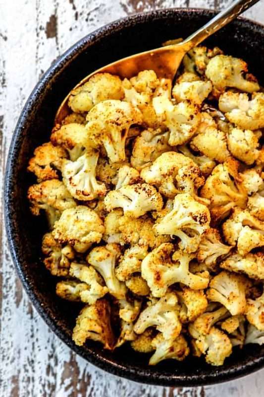 A large bowl of roasted cauliflower is prepared for serving with a metallic silver spoon.