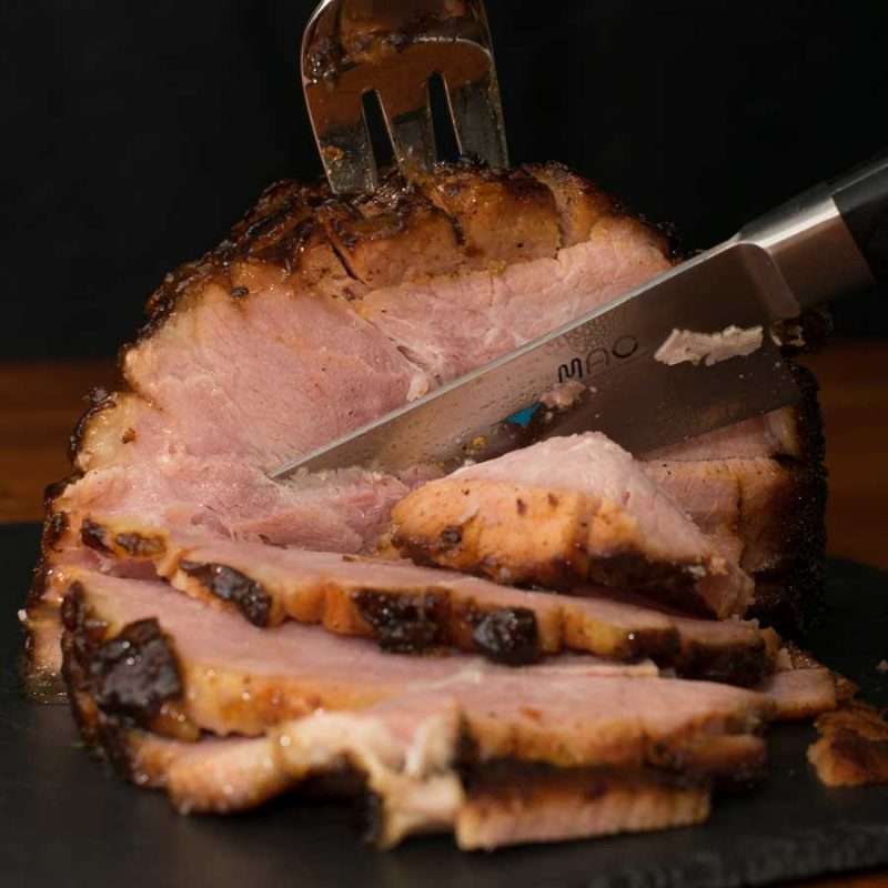 A fork and knife carve a juicy Maple Bourbon Glazed Grilled Ham into thick slices. This simple holiday recipe is made on a grill so the ham develops a crispy, caramel-colored crust on the edges. 