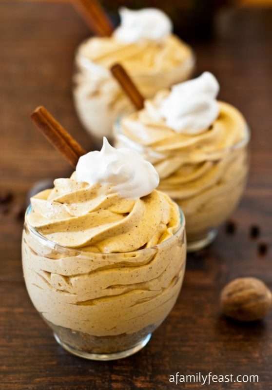 Small clear, glasses sitting on a wooden table are filled with No-Bake Mini Pumpkin Cheesecakes. Each mini cheesecake is topped with whipped cream and garnished with a cinnamon stick.   