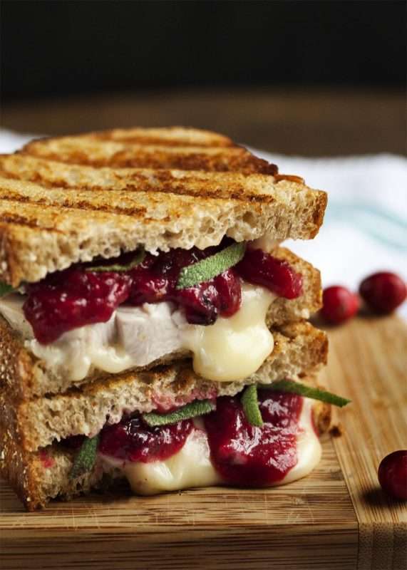 A panini sandwich with leftover thanksgiving turkey, melting brie cheese, and cranberry-mustard sauce sits on a wooden tabletop.