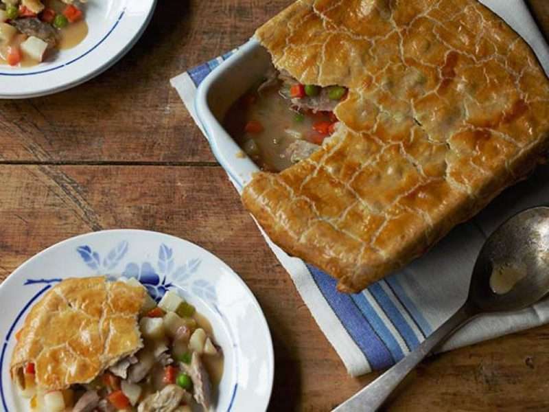 A turkey pot pie is served on a wooden table. There is a large casserole dish with the turkey pot pie on the right side of the image and two plates filled with portions of the turkey pot pie are on the left.