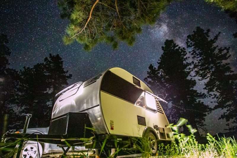 A teardrop trailer sits in the woods at night, surrounded by tall pine trees. There are thousands of stars visible in the dark, cloudless sky. 