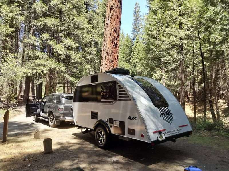 A teardrop travel trailer is towed by an SUV to a campsite in the woods. Tall trees surround the site.