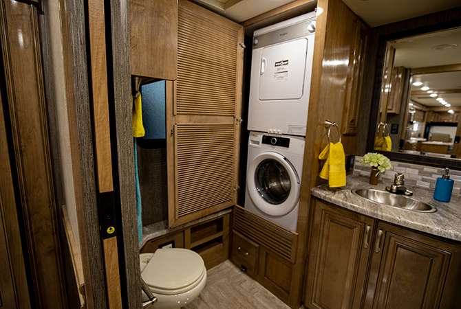 Inside the RV master bath of the Thor Aria 3901 is a porcelain toilet, stacked washer and dryer, and large vanity with mirror.