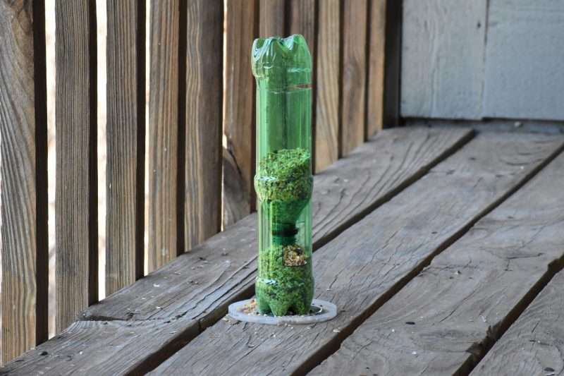 A DIY bird feeder, made from recycled soda bottles and a plastic lid sits o a balcony, filled with bird seed.