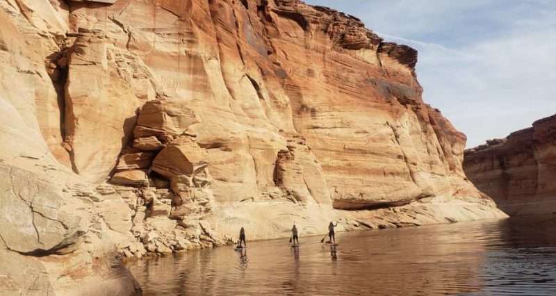 Three people on stand up paddleboards at Lake Powell in Utah. The paddleboarders are dwarfed by the massive rock wall behind them.