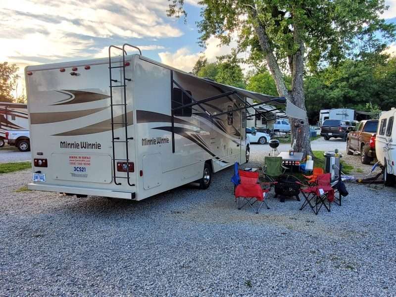 RVing tip: Rent before you buy. A rented Minnie Winnie Class C motorhome is parked at a busy campground. Several chairs are arranged around a fire pit across from to the RV.