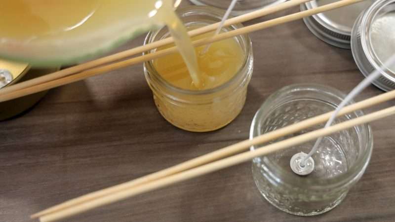 Melted wax is poured into a candle jar. A second empty jar sits nearby, ready to be filled. Bamboo skewers hold the wick of each candle in place during this process.