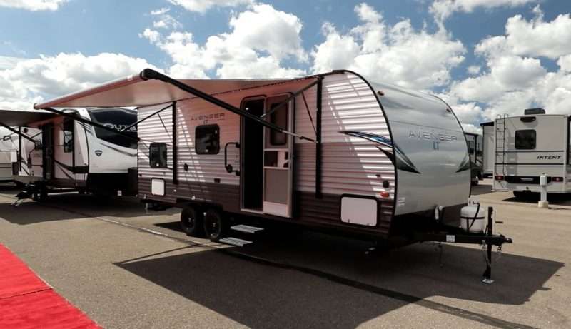 The Avenger LT 22BH travel trailer is parked on an RV dealership lot with its awning extended. It is a beautiful day with a blue sky and lots of fluffy white clouds.