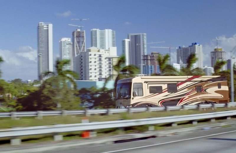 A Class A RV travels on a road lined with palm trees against a city skyscape in the background.