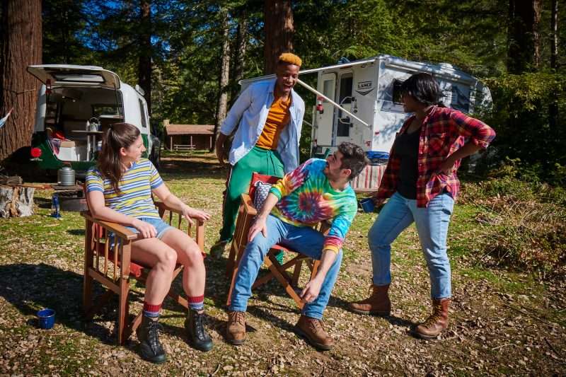 Four friends relax around a campsite where two small RVs are parked in the background.