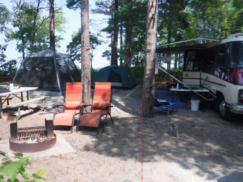 Twelvemile Beach Campground site 16 - showing a small RV and two tents on camp pad. Two red anti-gravity chairs sit next to a fire pit on the campsite.