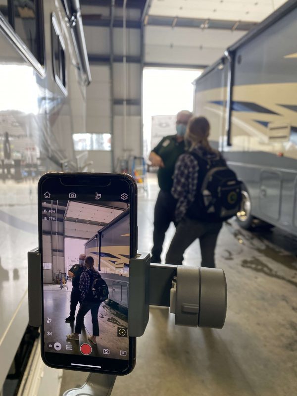 In the foreground, a smart phone is attached to a balancing device for filming. On the phone's screen and in the background, you can see what the recorder is recording: a young blond woman in a flannel shirt and faded black jeans wears a backpack while speaking to an older man with a dark green polo, black pants and blue face mask. They are standing next to a motorhome.