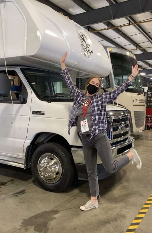 A young blond woman wearing a flannel shirt, faded black jeans and a black face mask, raises her arms and poses in front of a class C motorhome parked in a garage with the brand name Nexus on the front. 