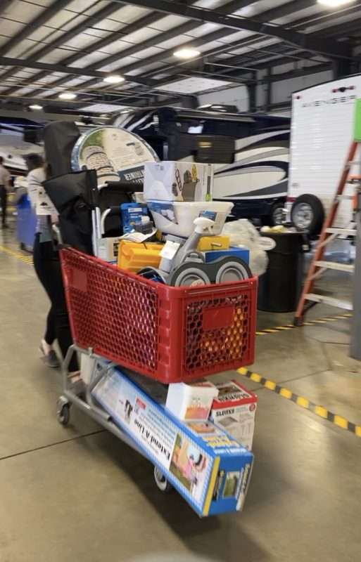 A red shopping cart filled with RV and camping accessories is pushed along a concrete floor in a large garage. There are multiple RVs in the background.