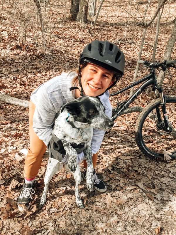 A young blond woman and a black and white dog pose in front of a black mountain bike on a forest trail covered with dry, fallen leaves.