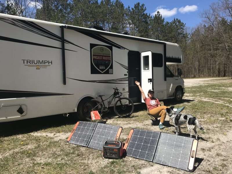 A young woman sits outside a Class C motorhome with her dog. She is sitting near her portable solar power setup which uses two large, folding solar panels to charge a lithium battery.