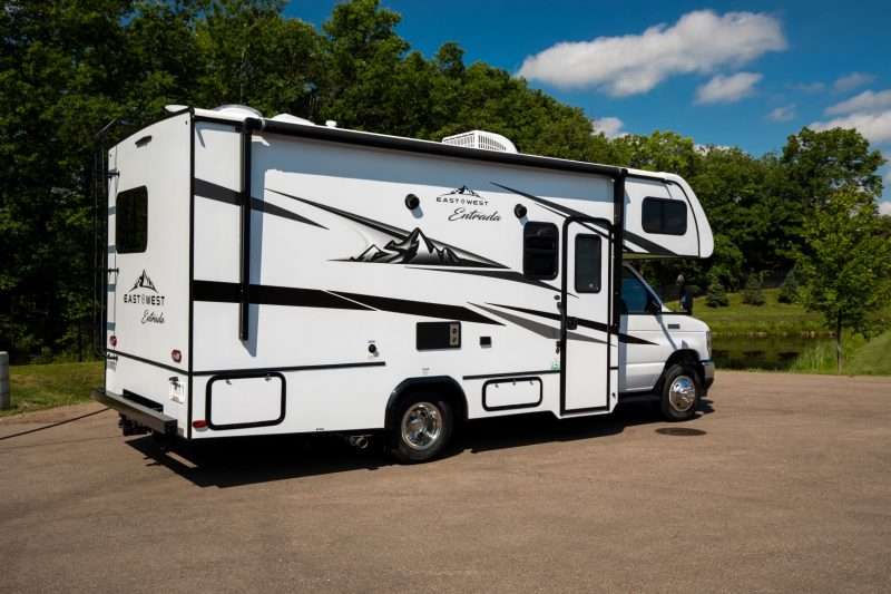 A white Class C motorhome with black and gray accent graphics is parked on a concrete pad near a pond and woods on a beautiful, blue-sky day.