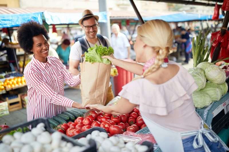 A couple purchases fresh fruits and vegetables from a woman at a farmers market.