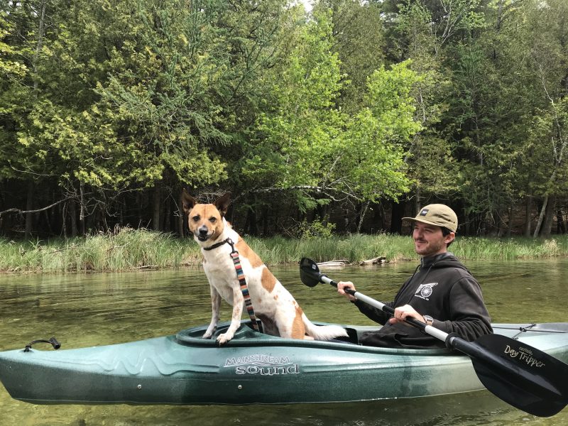 A man kayaks with his dog. The dog is standing up to look out over the water and towards the camera.