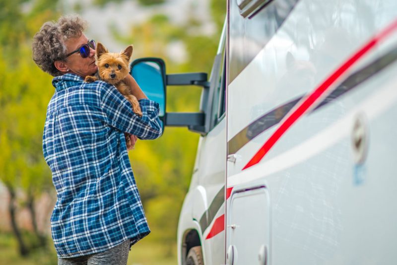 A woman carries her dog, a Yorkshire Terrier, in her arms as they stand outside an RV. She is wearing sunglasses and a plaid blue shirt. The dog is light brown and looking directly at the camera.