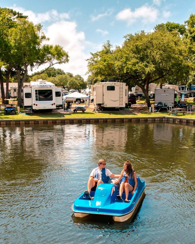 Two people use a paddleboat on the water while RVs are parked along the waterfront at Lakewood Camping Resort.