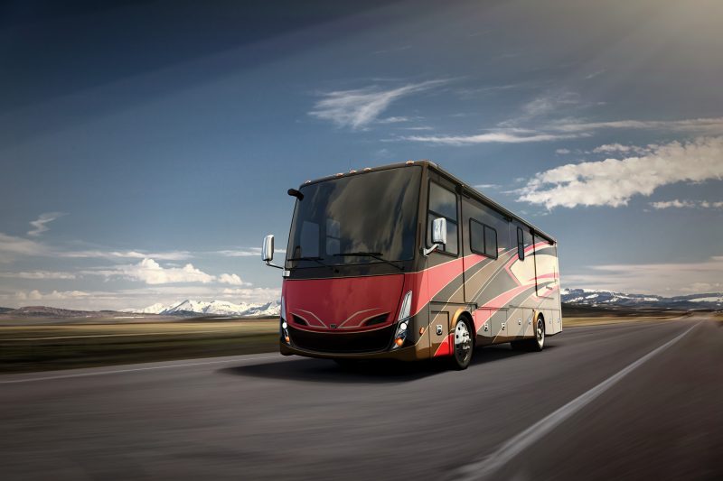 A modern Class A diesel RV with red, black, brown and tan colors travels down an open road.