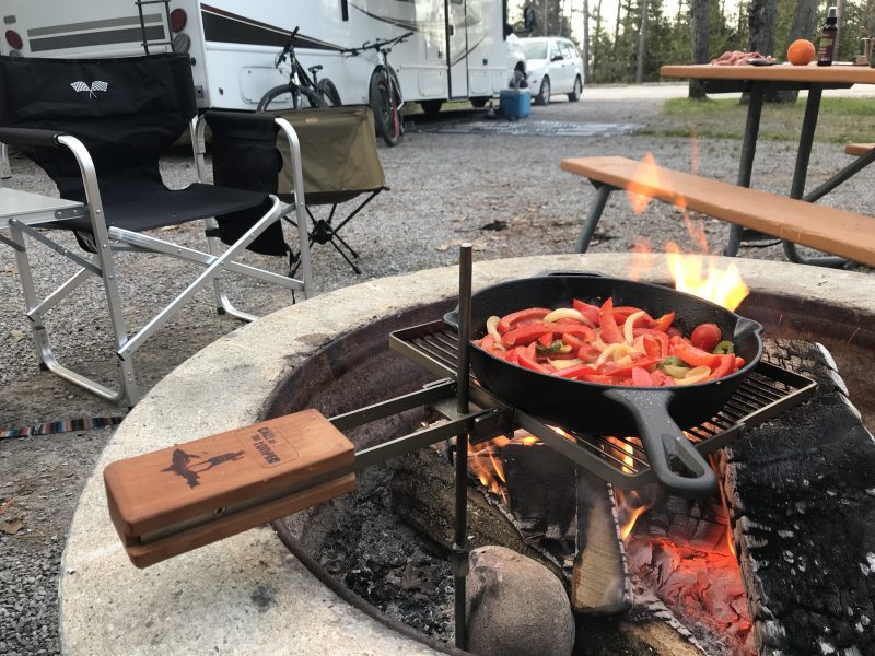 A cast iron skillet filled with chopped bell peppers and other vegetables cooks on a grill grate positioned over a campfire. In the background, an RV and vehicle are parked at a campsite. Two camp chairs are positioned near the fire.