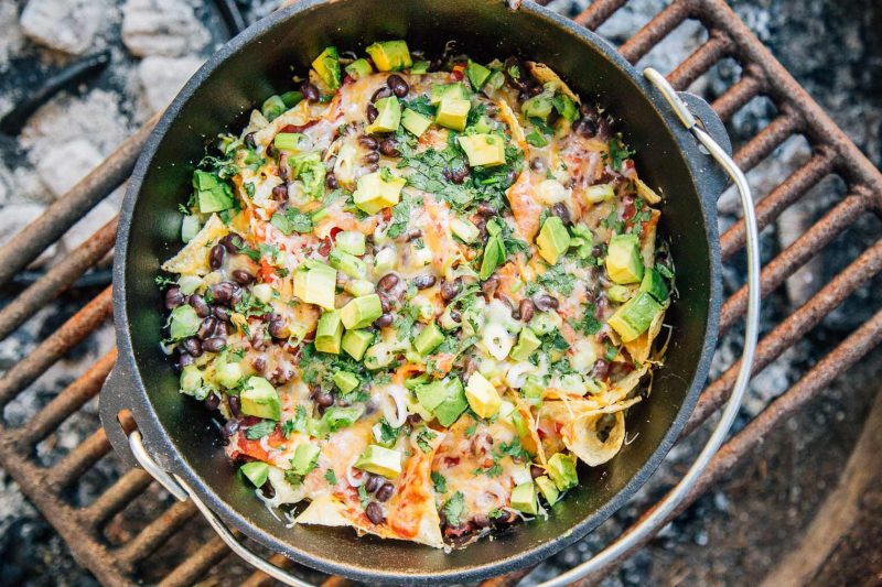A large cast iron pot sits on a grill grate over a campfire. The pot is filled with fixings for Campfire Nachos - tortilla chips are covered with beans, cilantro, melted cheese, avocado, onions, and more.