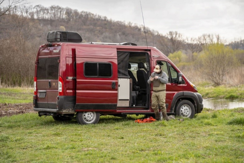 A man wearing waders and a hat stands outside a red Winnebago Solis Pocket preparing to go fly fishing. The RV is parked in a grassy, wooded area near a body of water. 