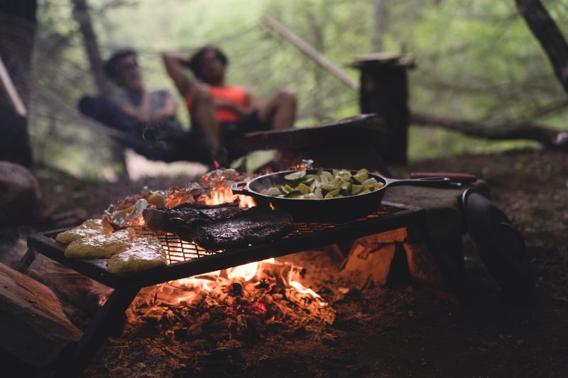 Two people in a hammock watch a fire with food cooking at a campsite.