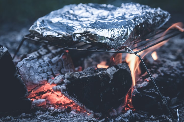 A pair of foil-packet meals cooks on a grill grate over a campfire. Foil packet meals are simple campfire recipes that are as easy as throwing all the ingredients into a piece of foil, folding the foil into a rectangular shape and cooking over a campfire.