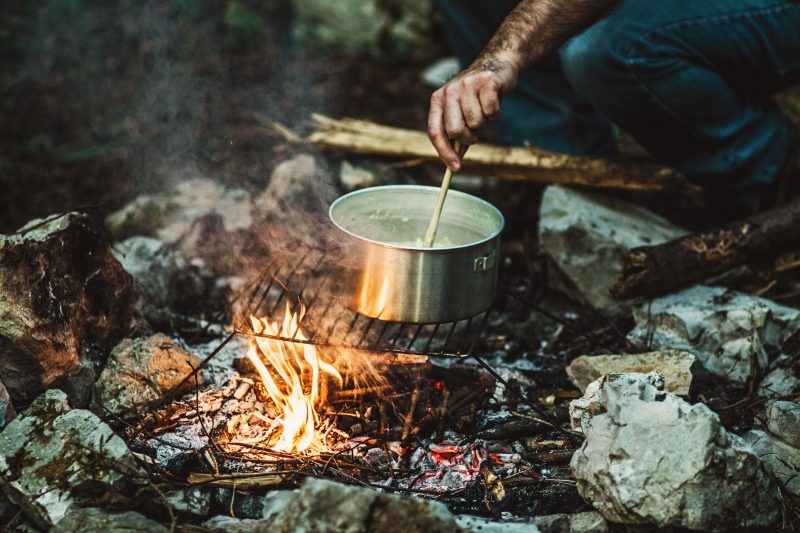 A man stirs a meal in a pot while it cooks over a campfire. One pot meals are ideal for camping cooking recipes because they mean less cleanup afterward.